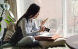 Woman with long dark hair sitting on a bed or couch next to a window looking at her phone to illustrate Holiday Deals Are Here. Don't Let Debt Follow